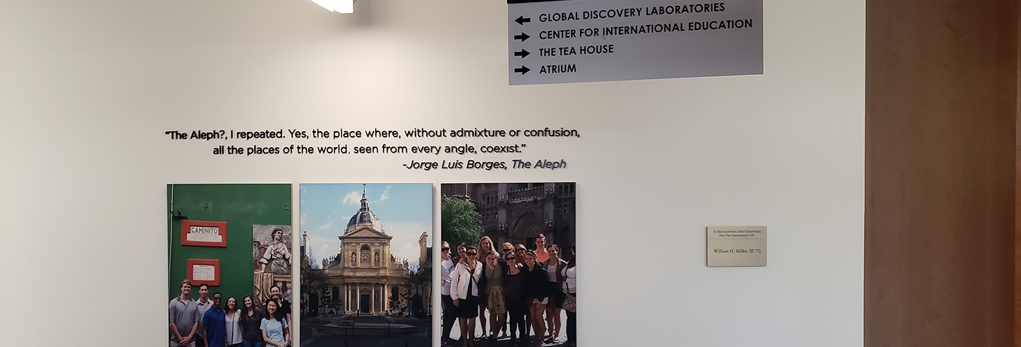 Printed photos of students studying abroad hang beneath a quote from Jorge Luis Borges' 'The Aleph', "The Aleph?, I repeated. Yes, the place where, without admixture or confusion, all the places of the world, seen from every angle, coexist." Above this, a sign points directs visitors to various departments and areas within the building.